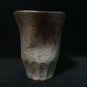Könnte beinhalten: Ein Keramikbecher mit einer strukturierten, unebenen Oberfläche, der am Rand einen Farbverlauf von Dunkelbraun zu Metallic-Bronze aufweist und nach unten hin in ein helleres, mattes Grau übergeht, mit vertikalen Einkerbungen am Boden.