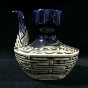May include: A ceramic pitcher with a dark blue top and spout, and a basket weave design on the base. The spout is curved and the base is a light gray color with dark gray accents. The pitcher is on a black surface.