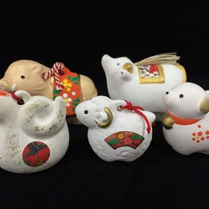 Könnte beinhalten: Eine Sammlung von fünf kleinen Tierfiguren aus Keramik, darunter ein beigefarbener Eber, ein weißer Ochse, eine weiße Schlange, ein weißes Schaf und ein weißer Hund. Jede Figur weist bemalte Details, florale Muster und kleine Schnüre auf.