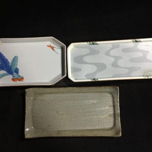 Könnte beinhalten: Drei rechteckige Serviertabletts. Ein weißes Tablett zeigt ein blaues Blattgemüse und einen kleinen orangefarbenen Vogel. Ein zweites weißes Tablett hat ein hellgraues Wellenmuster. Das dritte ist ein strukturiertes, gesprenkeltes beiges Keramiktablett.