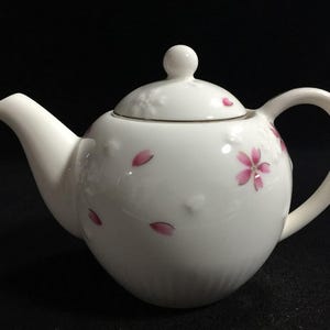 Peut inclure: Une théière en porcelaine blanche ornée d'un motif délicat de pétales de fleurs de cerisier roses dispersés sur la surface. La théière présente un corps arrondi, un bec verseur incurvé, un couvercle assorti avec un bouton et une finition lisse.