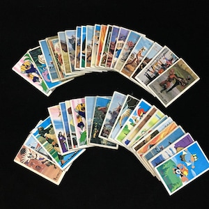 May include: A collection of vintage Japanese trading cards, arranged in two semi-circles against a black background. The cards feature colorful illustrations of various scenes and characters, with Japanese text.
