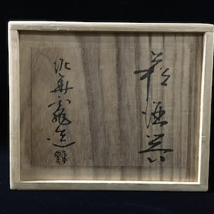 Può includere: Una scatola rettangolare in legno con venature color marrone chiaro e finitura liscia. Il coperchio presenta una calligrafia a inchiostro nero scritta in due colonne verticali, con un piccolo timbro rosso in fondo alla colonna di sinistra.