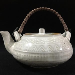 May include: A white ceramic teapot with a woven brown handle. The teapot features a decorative floral and geometric pattern. The spout and lid are also white. The teapot is a traditional design, suitable for serving tea.