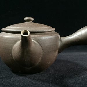 May include: A brown ceramic teapot with a rounded body, a curved handle, and a lid with a small knob. The spout is short and angled. The teapot is set against a dark background.