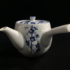 Op de afbeelding: Witte keramische theepot met een zijhandvat en een deksel. De theepot is versierd met blauwe bloempatronen. De tuit en het handvat zijn glad en gebogen. Het deksel heeft een kleine knop. De theepot is ontworpen voor theeservice.