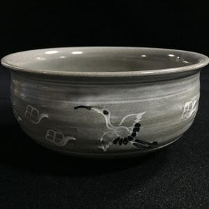 May include: A gray ceramic bowl with a hand-painted crane design. The bowl features a wide rim and a textured surface. The crane is depicted in white and black, with stylized details. Decorative elements include small, stylized house shapes.