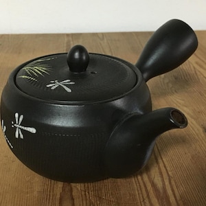 Peut inclure: Une théière kyusu en céramique noire mate avec une poignée latérale et un couvercle amovible. Le corps et le couvercle présentent des motifs de libellules peints en blanc et des accents d'herbe verte. La théière a un bec court et incurvé.