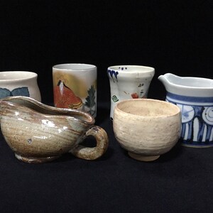 May include: A collection of six ceramic vessels including a beige gravy boat with a handle, a textured cream tea bowl, a white cup with a blue floral design, a cylindrical cup with a figure illustration, a white cup with blue accents, and a small bowl.