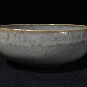May include: A round ceramic bowl with a speckled light gray and off-white glaze. The rim features a distinct brown, earthy tone that contrasts with the mottled, textured surface of the body, creating a simple and functional kitchen vessel.