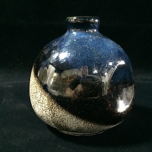 May include: A small, spherical ceramic vase with a narrow neck. The upper portion features a glossy, deep blue glaze that transitions into a textured, speckled beige and brown finish on the lower section, creating a distinct diagonal contrast.