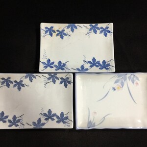 Puede incluir: Tres platos rectangulares de cerámica con fondo blanco. Dos platos presentan un patrón de hojas pintadas en azul en los bordes superior e inferior. El tercer plato muestra un delicado diseño floral en azul y rosa claro con tallos finos.