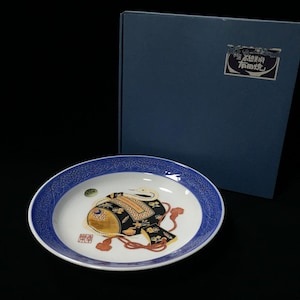 May include: A decorative ceramic plate with a blue patterned border and a central design featuring a black and gold motif. The plate is accompanied by a dark blue box with a label. The plate's design includes red accents.