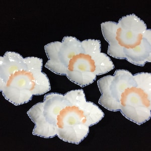 May include: A set of five white ceramic dishes shaped like maple leaves. Each dish features a central design with a pale yellow centre, a peach-coloured scalloped ring, and a light blue wash, finished with a dark blue painted edge detail.