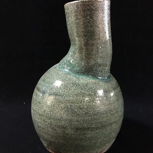 May include: A ceramic vase with a rounded base and an off-centre, elongated neck. The surface features a mottled, textured glaze in shades of sage green, olive, and dark grey, with a subtle horizontal banding pattern visible across the body.