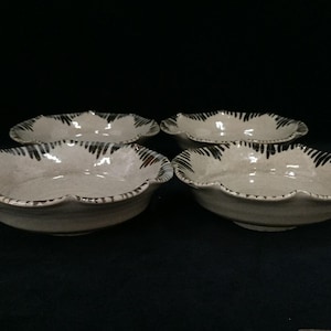 May include: A set of four shallow ceramic bowls with a light beige speckled glaze and a scalloped rim. Each bowl features dark brown, brush-stroke style markings along the edges, creating a repeating pattern around the entire circumference.