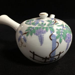 Könnte beinhalten: Eine weiße Keramik-Teekanne mit seitlichem Griff und einem Deckel, der mit einem bemalten Motiv aus violetten Glyzinienblüten, grünen Blättern und blauen Bambusstangen verziert ist. Der Boden der Kanne zeigt ein aufgemaltes braunes Zaunmotiv.