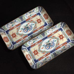 May include: Two rectangular ceramic serving trays with ornate floral and bird designs. The trays feature a cream-colored background with red, blue, and green accents. The edges are trimmed with gold.
