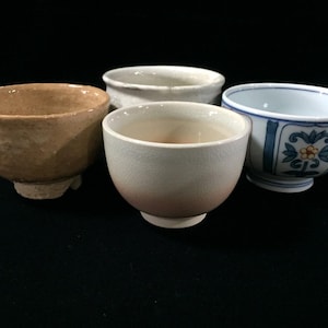 May include: A collection of four ceramic bowls featuring distinct designs. One bowl is textured tan, one is off-white with a subtle gradient, one is plain white, and one is white with blue and orange floral patterns and blue vertical stripes.