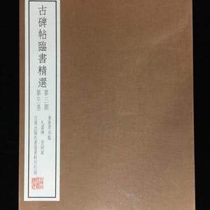 May include: A rectangular brown book with a white vertical label on the left side. The label features black Chinese characters reading 古碑帖臨書精選, 第三期, 第十一卷, 桑原翠邦臨, 禮器碑, 曹全銘, and 日寶出版社書道資料刊行部, along with a small red square seal at the bottom.