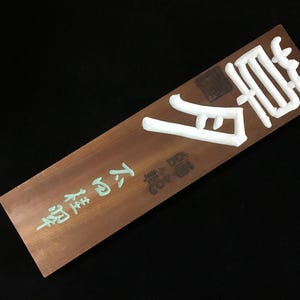 Könnte beinhalten: Eine rechteckige Holztafel mit glatter brauner Oberfläche, die oben große, erhabene weiße japanische Schriftzeichen aufweist. Darunter sind kleinere hellgrüne japanische Schriftzeichen eingraviert, ergänzt durch einen dunklen quadratischen Stempel.