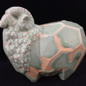 Könnte beinhalten: Eine Keramikfigur in Form eines Schafs mit facettiertem, geometrischem Körper, einer hellgrünen Glasur und sichtbaren pfirsichfarbenen Tonkanten. Der Kopf- und Halsbereich ist strukturiert und lockig, während der Körper glatte, kantige Flächen aufweist.