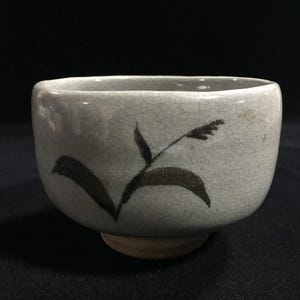 May include: A ceramic tea bowl with a pale grey glaze and a dark grey floral design. The bowl has a wide, rounded shape and a small, unglazed foot. The floral design is simple and elegant, adding a touch of nature to the piece.