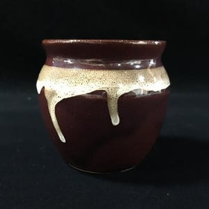 May include: A small, rounded ceramic pot with a glossy dark brown finish. The upper portion features a contrasting band of speckled cream-colored glaze that drips down the sides of the vessel, creating a textured, layered appearance against the dark base.