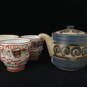 Può includere: Una teiera in ceramica con base beige, fasce orizzontali blu e motivi a spirale marrone scuro, accompagnata da due tazze abbinate con volute floreali rosse e verdi e un'etichetta bianca con testo giapponese sulla parte anteriore.