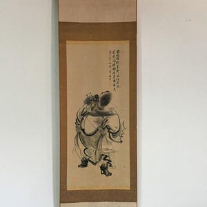May include: A traditional Asian scroll painting featuring a black and white ink illustration of a figure with a long beard and flowing robes. The artwork is mounted on a tan and brown scroll with a black rod at the bottom.