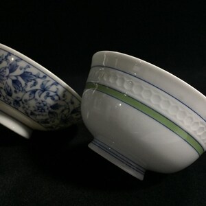 May include: Two ceramic bowls with distinct designs. One bowl features a blue and white floral pattern covering the exterior. The second bowl is white with a green and blue horizontal stripe and a textured honeycomb pattern around the upper rim.