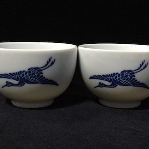 May include: Two white ceramic tea cups with a blue crane design. The crane is depicted in flight, with its wings spread. The cups have a rounded shape and sit on a dark surface. The cups are likely part of a tea set.