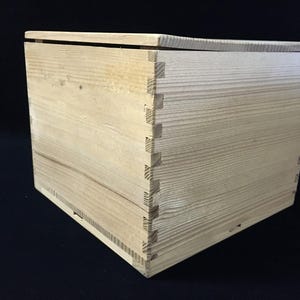 May include: A rectangular wooden box with a light tan natural finish, featuring visible wood grain patterns and interlocking finger joints at the corners. The box includes a flat, matching wooden lid that sits flush on top of the structure.