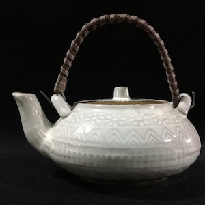 Op de afbeelding: Witte keramische theepot met een bruin geweven handvat. De theepot heeft een bloemen- en geometrisch reliëfpatroon rond de body. De tuit en het deksel zijn ook wit, wat een samenhangend ontwerp creëert. Een klassieker voor theeliefhebbers.