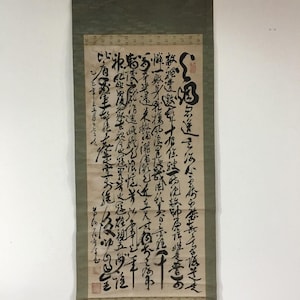 May include: A vertical hanging scroll featuring black ink calligraphy on a light beige paper background. The scroll is mounted on a muted olive green fabric border with a wooden rod at the top and bottom. Red ink stamps are visible on the paper.