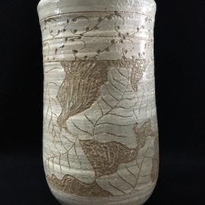 May include: A cylindrical ceramic vase with a light beige and cream glaze. The surface features a textured, carved pattern of large leaves and a delicate vine motif near the rim. The finish is matte in the carved areas and glossy elsewhere.