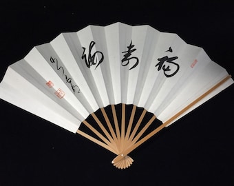 Puede incluir: Un abanico plegable blanco con varillas de madera de color marrón claro. El abanico presenta caracteres de caligrafía japonesa en negro sobre la superficie y dos pequeños sellos cuadrados rojos en el lado izquierdo. Está abierto en semicírculo.