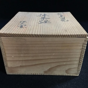 Può includere: Una scatola di legno color chiaro con coperchio. La scatola ha venature del legno visibili e sembra fatta a mano. Caratteri giapponesi sono stampati sul coperchio. La scatola è rettangolare e poggia su una superficie scura.