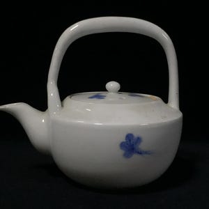 Peut inclure: Une théière en céramique blanche dotée d'une anse haute et arquée ainsi que d'un bec court et pointu. Le corps et le couvercle sont ornés de petits motifs floraux bleus peints à la main. La théière présente une forme arrondie et trapue.