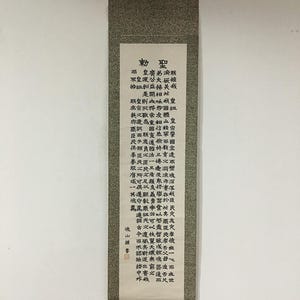 May include: A vertical wall hanging scroll featuring a long piece of off-white paper with dense black Chinese calligraphy characters arranged in columns. The scroll is framed by a textured grey fabric border and includes a small red seal stamp.