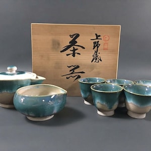 Op de afbeelding: Een keramische theeset bestaande uit een theepot met deksel, een kleine kom en vijf kopjes. Elk stuk heeft een crèmekleurige basis en een levendig blauwgroen glazuur aan de bovenzijde. Achter de set staat een houten doos met zwarte kalligrafie.