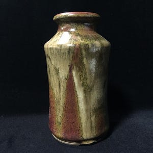 Puede incluir: Un jarrón cilíndrico de cerámica con cuello estrecho y borde ensanchado. La superficie presenta un esmalte reactivo con goteos verticales en tonos crema, verde oliva y marrón rojizo intenso, creando un aspecto texturizado y multicolor.