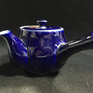 Peut inclure: Une théière en céramique bleu cobalt profond avec une poignée latérale et un couvercle assorti. La théière présente une finition brillante et un petit bec verseur. Un petit motif floral coloré est visible sur le dessus du couvercle.