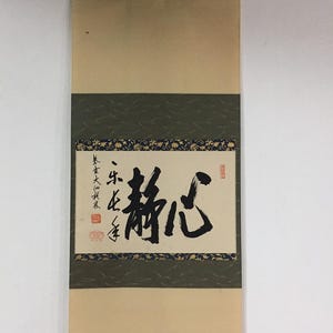 Può includere: Un rotolo di pittura giapponese tradizionale con calligrafia a inchiostro nero. Il rotolo presenta uno sfondo beige, una fascia verde scuro con un motivo floreale e un'asta di legno nera nella parte inferiore. La calligrafia include i caratteri "静心".