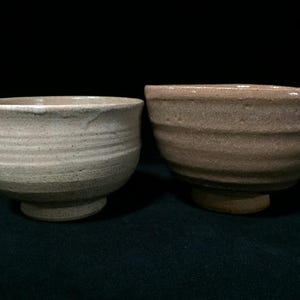 May include: Two ceramic bowls with a textured, ribbed exterior. The bowl on the left is a light cream color with subtle speckles, while the bowl on the right is a muted, earthy tan color. Both bowls feature a wide rim and a raised circular base.