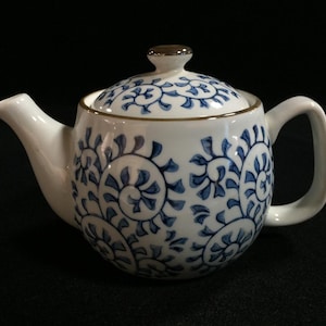 Könnte beinhalten: Weiße Keramik-Teekanne mit blauem Blumenmuster. Die Teekanne hat einen gebogenen Ausguss und einen Henkel. Der Deckel ist mit einem kleinen, goldfarbenen Knopf versehen. Das Design ist klassisch und eignet sich zum Servieren von Tee.