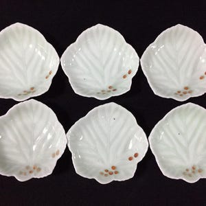 May include: Six white leaf-shaped dishes with a delicate, pale green leaf vein pattern. Each dish has small, brown, circular accents near the base. The dishes are arranged on a dark surface.