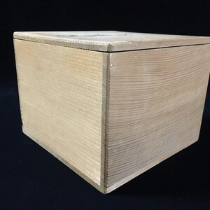May include: A square wooden box with a light tan, natural wood grain finish. The box features a flat, removable lid that sits flush with the top edges. The construction shows visible vertical wood grain patterns on the sides and a smooth surface.