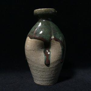 May include: A handcrafted ceramic vase with a unique, organic shape. The upper portion is glazed in a deep green, with streaks of brown, flowing over a textured, beige lower half. The vase has a narrow neck and a dark green rim.