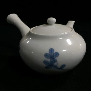 Może przedstawiać: Biały ceramiczny czajniczek z pokrywką i uchwytem. Czajniczek ma niebieski kwiatowy wzór na korpusie. Wylewka i uchwyt są zakrzywione. Pokrywka ma mały guziczek. Czajniczek jest przeznaczony do parzenia herbaty.